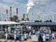Nigerian engineers in lab coats work with advanced machinery in a modern industrial facility, surrounded by solar panels, wind turbines, and rising smokestacks under a bright sky.