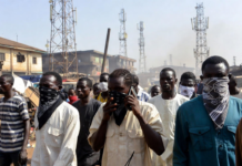 Bandits Bounce Calls Off Multiple Towers – Minister Masked individuals using mobile phones in front of multiple telecom towers in a dusty Nigerian rural area, illustrating bandits evading security forces by routing calls through different base stations.