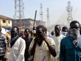Bandits Bounce Calls Off Multiple Towers – Minister Masked individuals using mobile phones in front of multiple telecom towers in a dusty Nigerian rural area, illustrating bandits evading security forces by routing calls through different base stations.