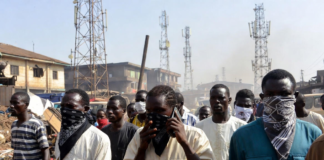 Bandits Bounce Calls Off Multiple Towers – Minister Masked individuals using mobile phones in front of multiple telecom towers in a dusty Nigerian rural area, illustrating bandits evading security forces by routing calls through different base stations.