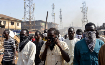 Masked individuals using mobile phones in front of multiple telecom towers in a dusty Nigerian rural area, illustrating bandits evading security forces by routing calls through different base stations.
