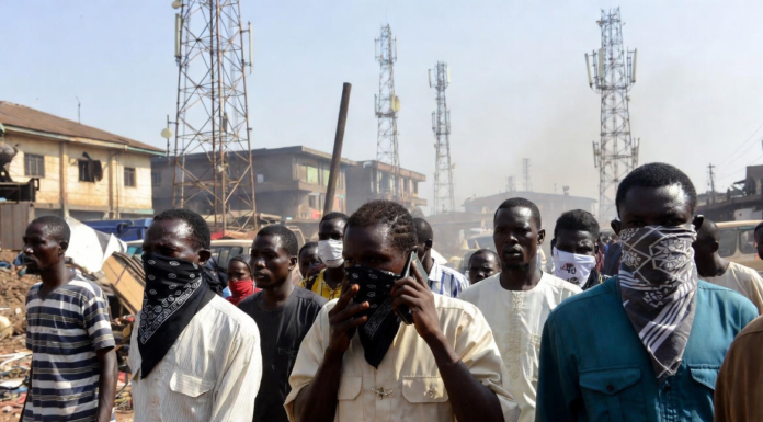Bandits Bounce Calls Off Multiple Towers – Minister Masked individuals using mobile phones in front of multiple telecom towers in a dusty Nigerian rural area, illustrating bandits evading security forces by routing calls through different base stations.