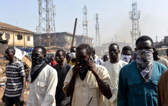Minister Reveals Bandits Are Bouncing Calls Off Multiple Telecom Towers to Evade Tracking Masked individuals using mobile phones in front of multiple telecom towers in a dusty Nigerian rural area, illustrating bandits evading security forces by routing calls through different base stations.