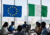 EU Commits €45 Million to Boost Nigeria’s Digital Economy Nigerian and European officials in a modern conference room with EU and Nigerian flags, collaborating on laptops against a Lagos city skyline, symbolising partnership in digital innovation.