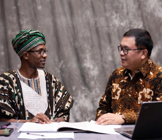 Nigerian official in traditional attire and Indonesian livestock expert reviewing digital tablet together, with background overlays of modern cattle farms, AI monitoring systems and dairy processing plants.
