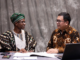 Nigerian official in traditional attire and Indonesian livestock expert reviewing digital tablet together, with background overlays of modern cattle farms, AI monitoring systems and dairy processing plants.